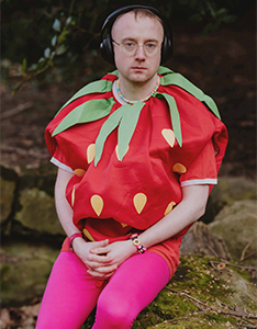 Der Komponist Alex Paxton im Erdbeerkostüm sitzt mit Kopfhörern auf einem Felsen im Freien.