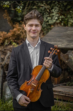 Der Geiger David Hernych steht mit Instrument Im Anzug vor Bäumen und einer Holzhütte