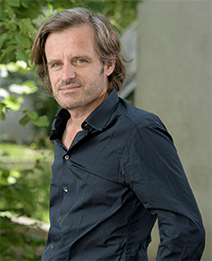 Porträt von Andri Hardmeier mit mittellangen Haaren in dunklem Hemd. Er steht im Freien vor grünem Laub und blickt mit ernstem Gesichtsausdruck in die Kamera.