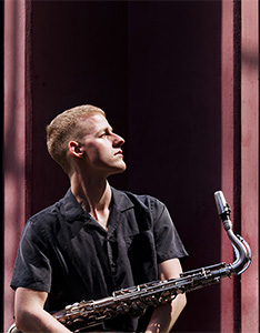 Fabian Willmann (sitzt mit Saxofon vor grossen Fenstern) wird ab Herbstsemester 2026/27 Dozent für Jazz-Saxofon an der HSLU Musik. 