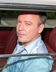 Ein Mann im hellblauen Hemd sitzt auf einem Rücksitz im Auto und blickt aus dem offenen Fenster.