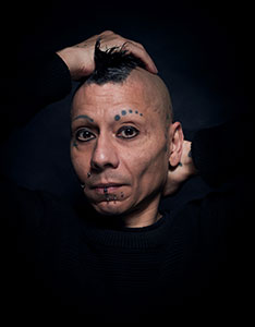 Porträt von Cedrik Fermont mit Irokesenschnitt, Gesichtspiercings und Tätowierungen auf der Stirn, die beide Hände hinter dem Kopf verschränkt hat. 