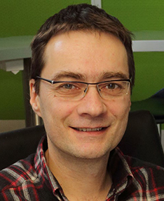 Porträt von Walter Fölmli mit kurzen braunen Haaren, Brille und kariertem Hemd. Er lächelt freundlich und sitzt in einem modernen Büroumfeld mit grüner Wand und schwarzen Stühlen im Hintergrund. Bild: zvg