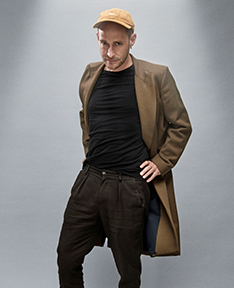 Portrait von Andreas Schaerer mit Kappe, dunklem Outfit und Mantel vor grauem Hintergrund, in lässiger Pose. Bild: Gregor Hohenberg