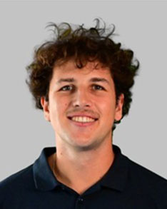 Smiling man with curly brown hair wearing a dark polo shirt against a neutral background.