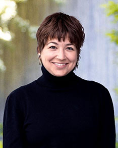 Smiling person with short brown hair wearing a black turtleneck, photographed outdoors with a blurred background.