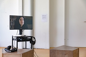 Eine Ausstellungssituation mit einem Bildschirm auf dem eine Frau zu sehen ist, und zwei Kopfhörern und zwei Hockern