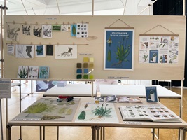 Eine Ausstellungstafel und verschiedene Illustrationen über Wildkräuter, insbesondere Spitzwegerich, Löwenzahn, Brennnessel und Bärlauch. Die Gestaltung kombiniert detailreiche Schwarz-Weiss-Skizzen und farbige Illustrationen, ergänzt durch erklärende Texte. Im Vordergrund ein Faltheft in der Hand, das mit klaren Grafiken den praktischen Gebrauch der Pflanzen darstellt.