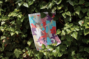 Buntes Plakat im Gebüsch – Sommerplakat mit verspieltem, pflanzenähnlichem Muster in kräftigen Farben, halb im Gebüsch platziert.