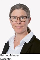 Portrait von Bettina Minder. Bettina hat dunkelblonde Haare, die sie in einem tiefen Zopf zusammengebunden hat. Sie trägt eine Brille und ein weisses Hemd mit schwarzem Blaser.