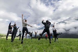 Eine Gruppe junger Menschen tanzt in der Natur auf einer Wiese. Der Himmel ist bewölkt. 
