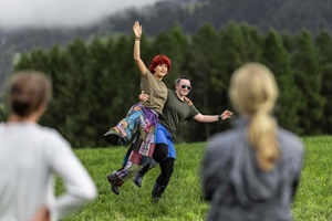 Zwei junge Menschen tanzen in der Natur auf einer Wiese. Im Vordergrund sind weitere Menschen von hinten erkennbar. 