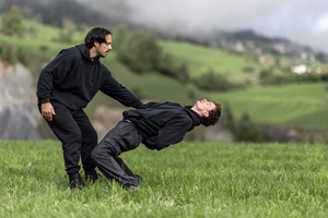 Zwei junge Menschen tanzen in der Natur auf einer Wiese. Im Hintergrund sind Felder und Häuser erkennbar. Der Himmel ist bewölkt. 