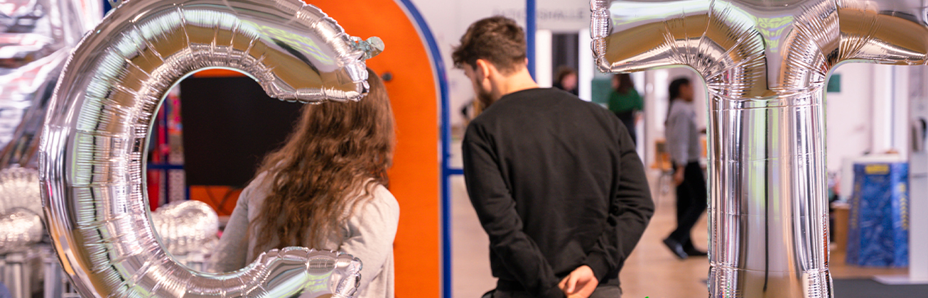 Zwei Personen stehen in einem Ausstellungsraum, eingerahmt von silbernen Ballonbuchstaben „C“ und „T“ – freundliche Atmosphäre beim Eintreten in eine kreative Umgebung.