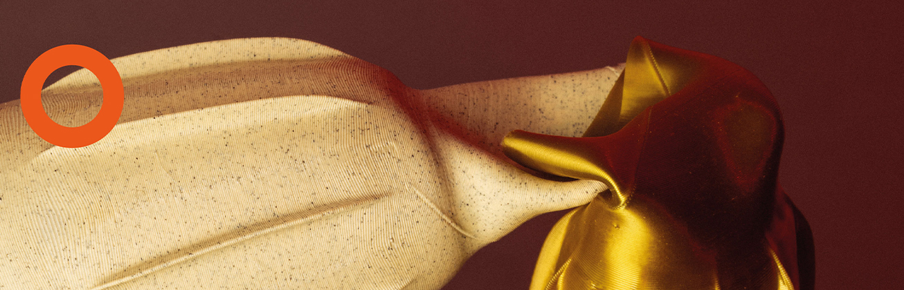     Nahaufnahme eines hellen, fein gesprenkelten Textils mit Falten, das rechts mit glänzendem gold- und dunkelrotem Stoff verknotet ist.