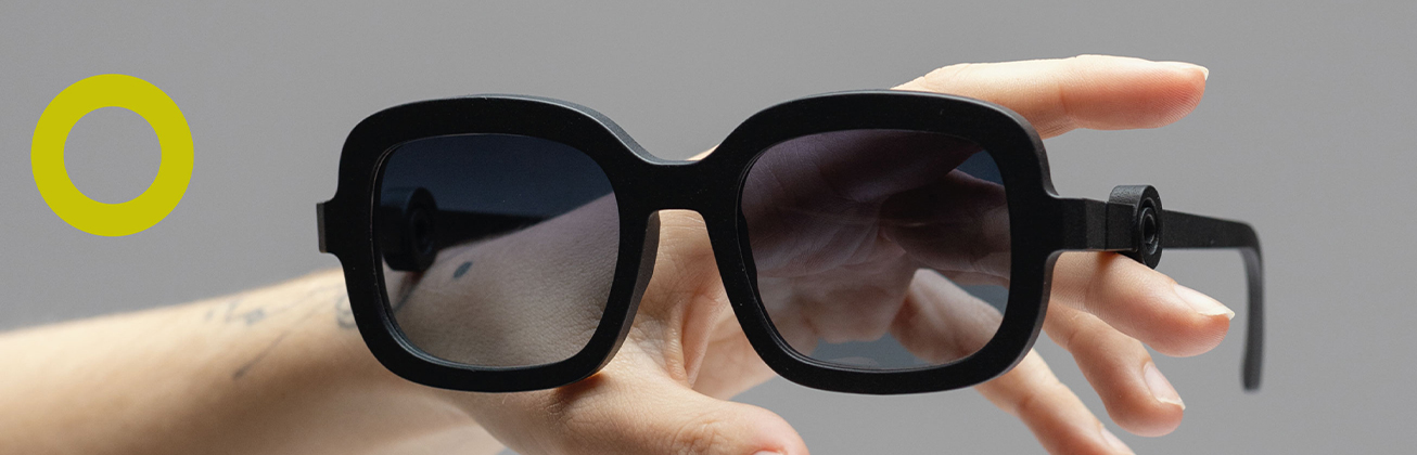     Nahaufnahme einer schwarzen Sonnenbrille mit breitem, eckigem Rahmen und dunklen Gläsern, die von einer Hand vor grauem Hintergrund gehalten wird.
