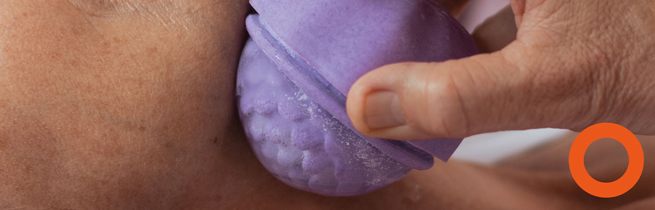     Nahaufnahme einer Hand, die ein rundes violettes Objekt mit transparenter Struktur an die nackte Haut einer Person drückt.