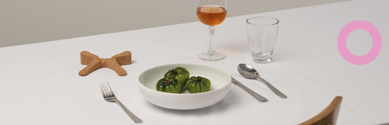     Weiss gedeckter Tisch mit einem Teller mit drei grünen Tomaten, Besteck, zwei Gläsern und einem kleinen braunen Objekt in Schleifenform.