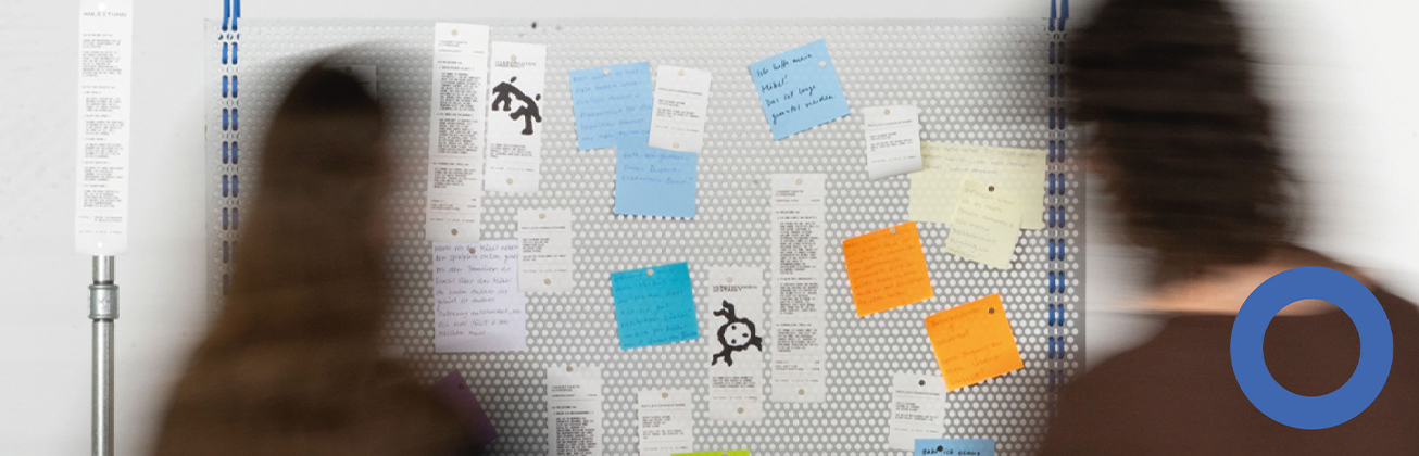     Metallische Lochwand mit zahlreichen weissen Zetteln und farbigen Haftnotizen in Blau, Orange und Gelb; davor bewegen sich unscharf zwei Personen.