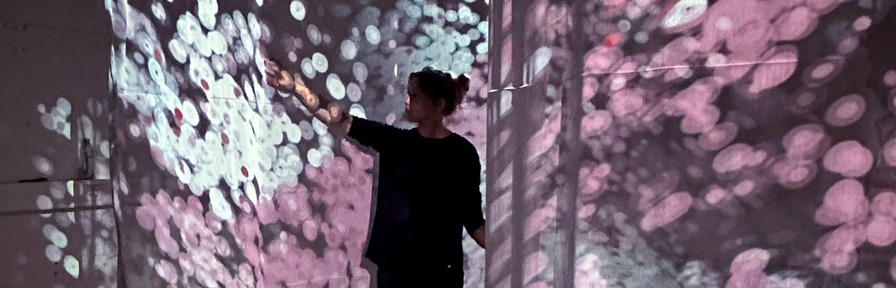 Eine Frau steht in der Mitte einer Projektion von weissen und rosafarbenen verschiedengrossen Punkten