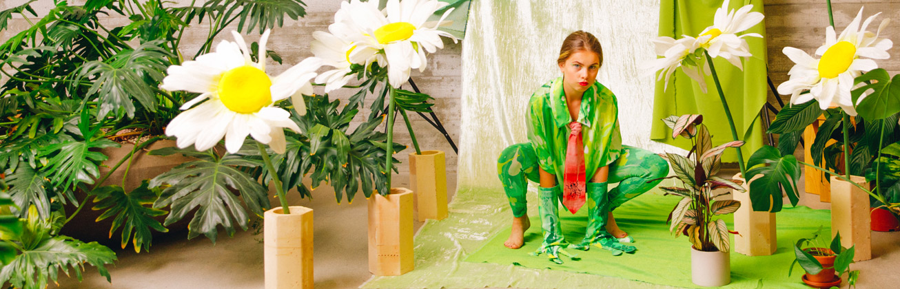 Studentin, die vor einer Leinwand und umzingelt von Blumen und Pflanzen auf dem Boden kauert und grün gekleidet ist, schaut in die Kamera