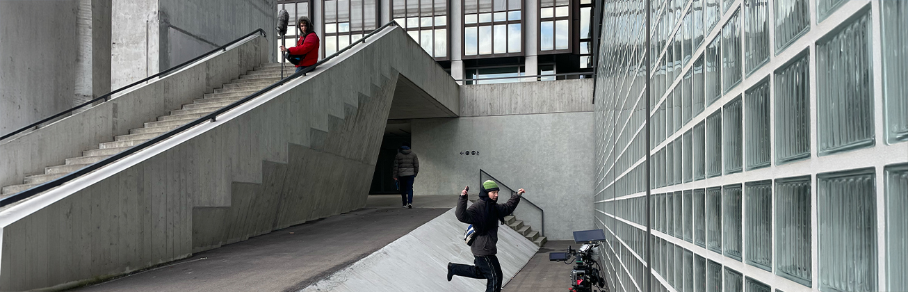 Eine Betontreppe und viele Fenste, eine Person in schwarz gekleidet mit einer Kamera und eine Person auf der Treppe, die in rot gekleidet ist. 