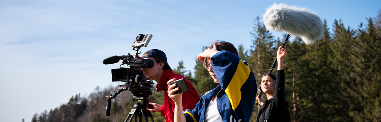 Eine Naturszene mit blauem Himmel. Man sieht drei Personen, eine filmt die Natur, die andere nimmt den Ton auf und die dritte Person schaut sich das Bild auf dem Handy an. 