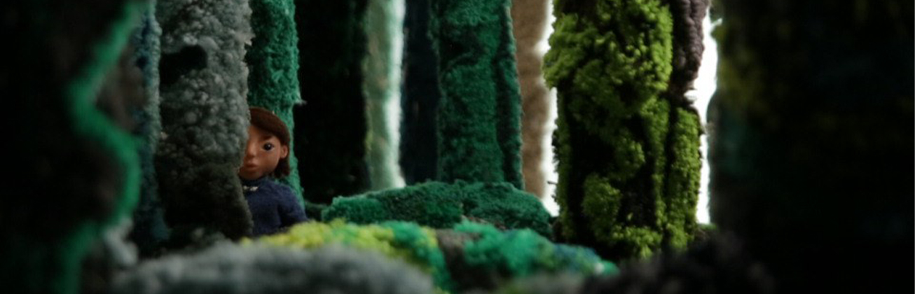     Papo and Pepe: Eine kleine animierte Figur blickt zwischen hohen, dicht mit grünem und grauem Garn bedeckten Baumstämmen hervor. Die textile Landschaft erinnert an einen üppigen Wald.