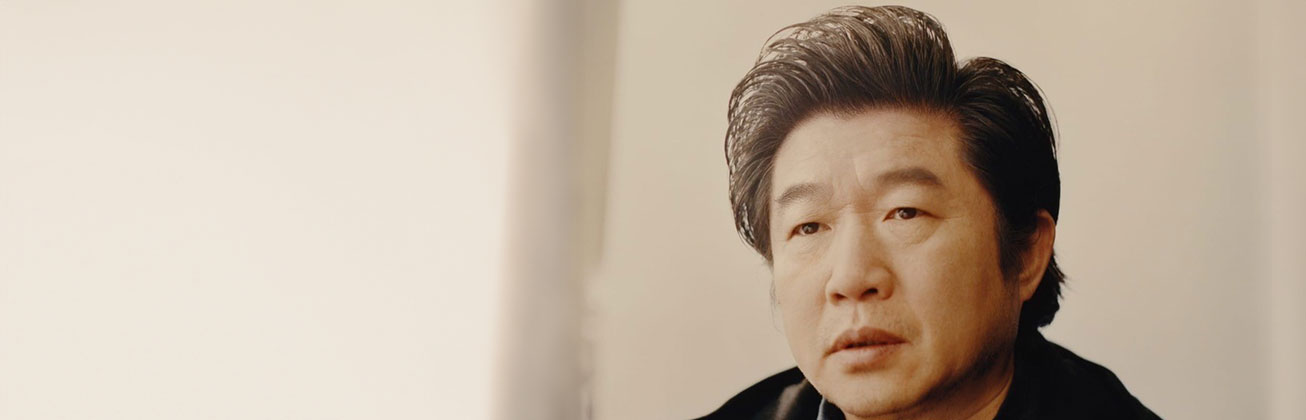 Portrait von Wang Bing, der ernst in die Kamera schaut, eine weisse Wand ist im Hintergrund