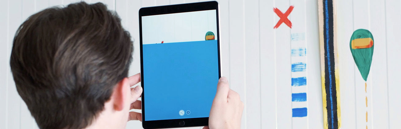 Ein Student richtet sein iPad auf eine Wand mit Zeichnungen, die auf dem BIldschirm anders dargestellt übertragen werden