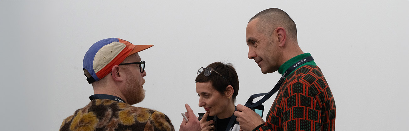 Zwei Männer und eine Frau in der Mitte, die farbig gekleidet sind und vor einer weissen Wand diskutieren 