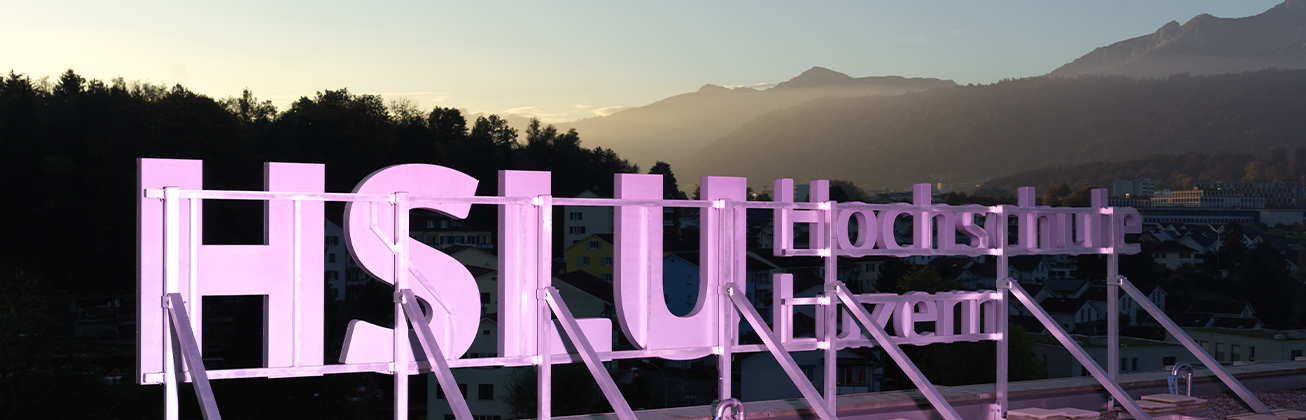 Leuchtschrift der Hochschule Luzern – HSLU Design Film Kunst mit Blick über Luzern und die Zentralschweizer Alpen bei Sonnenaufgang.