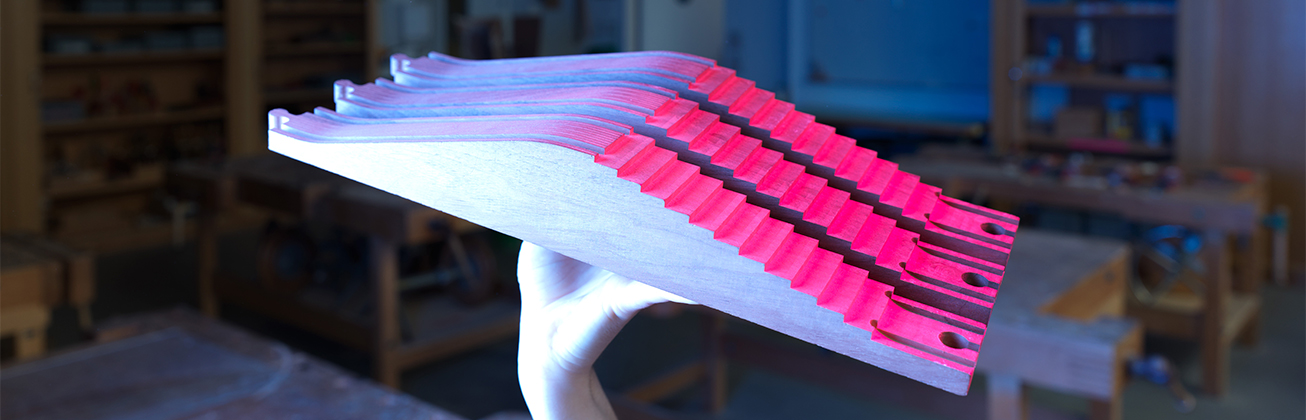 Mehrschichtiges, gefrästes Holzobjekt mit wellenförmigem Profil und pink markierten Stufen, von einer Hand gehalten in einer Werkstattumgebung mit unscharfem Hintergrund, als Beispiel für experimentelle Holzverarbeitung und präzise Handwerksarbeit.