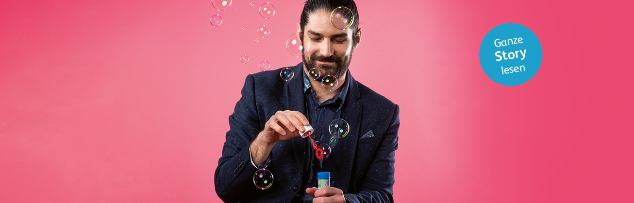 Ein Mann mit Bart befindet sich in der Mitte vor einer pinken Wand und ist damit beschäftigt, Seifenblasen zu produzieren