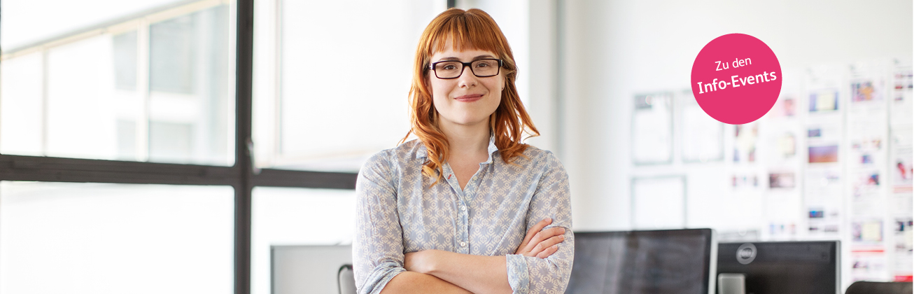 Eine rothaarige Frau steht mit verschränkten Armen in der Mitte einer weisslichen Büroräumlichkeit und lächelt in die Kamera