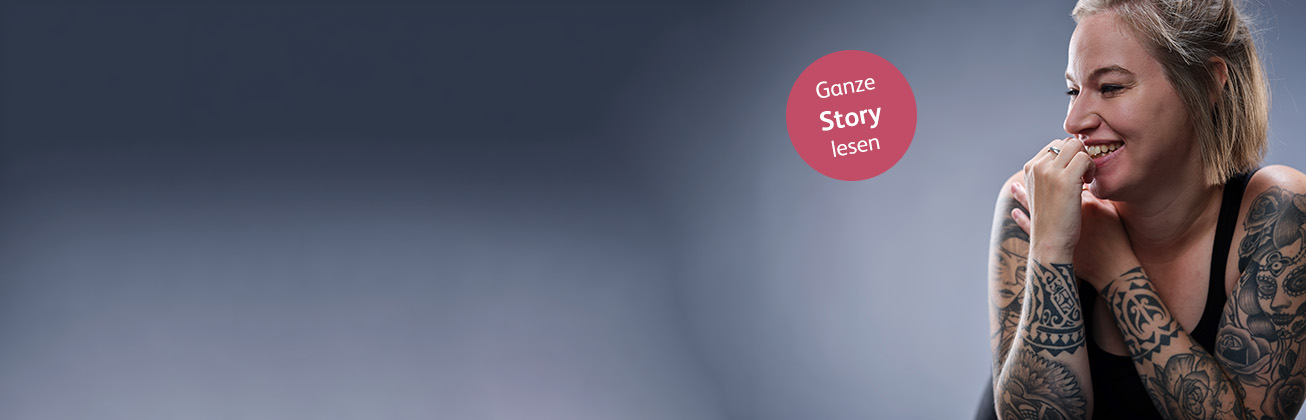 Eine blonde tättowierte Frau auf der rechten Seite des Bildes lächelt nach Links und der Hiintergrund besteht aus weichen Grautönen