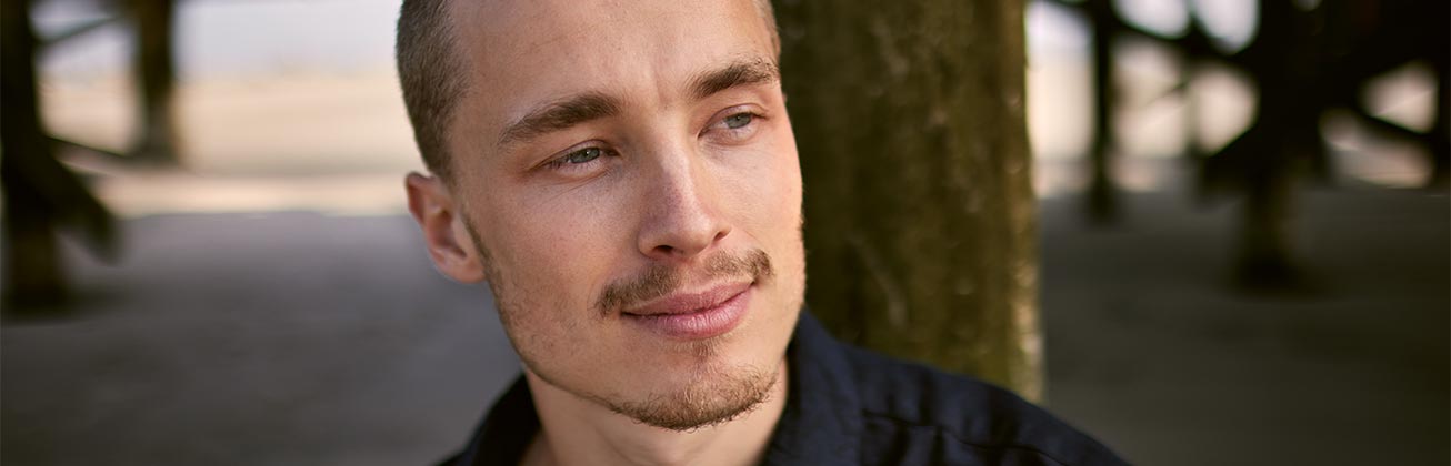 Junger Mann blickt zuversichtlich in die Ferne und im Hintergrund sind Baumstämme zu sehen 