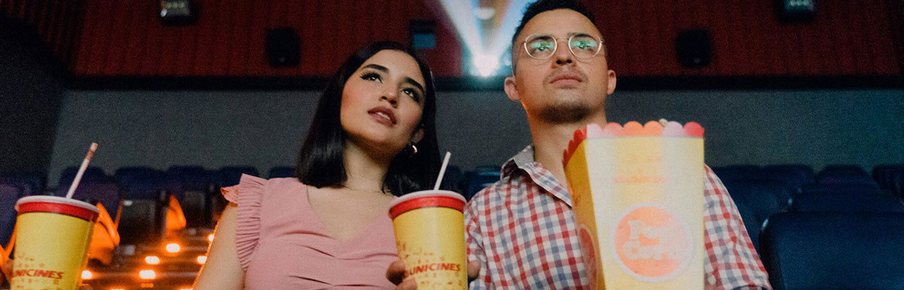 Eine Frau und ein Mann sitzen nebeneinander im Kino und halten Getränke und Popcorn in der Hand