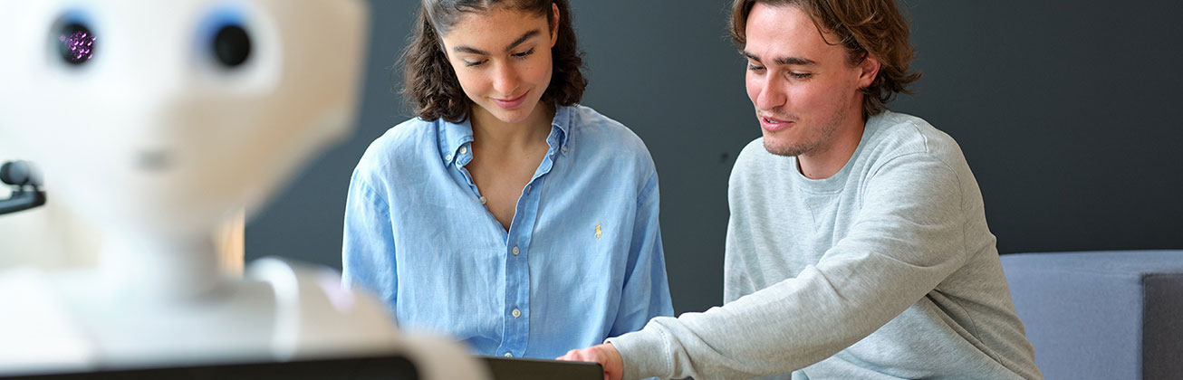 Eine Frau und ein Mann arbeiten lächelnd zusammen und links im Vordergrund steht ein Roboter