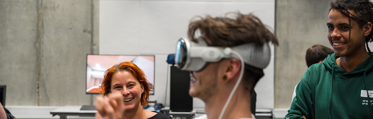 Ein junger Mann trägt eine VR-Brille, eine Frau und ein Mann im Hintergrund lachen