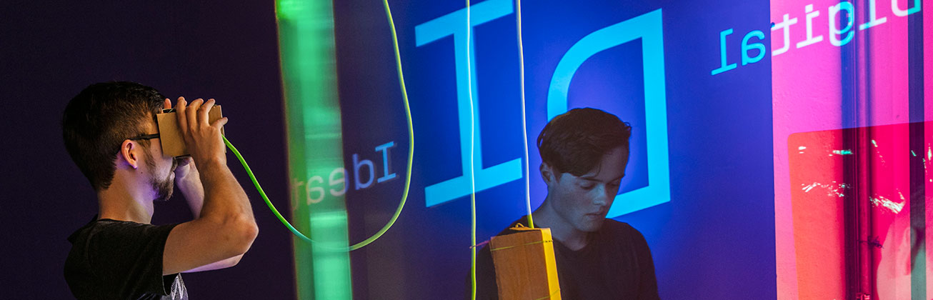 Ein Student mit Virtual-Reality-Brille interagiert mit einer digitalen Umgebung, während ein ander Student vor bunten, reflektierenden Glasflächen mit digitalen Schriftzügen steht