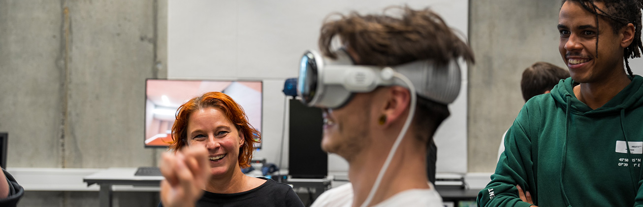 Ein junger Mann trägt eine VR-Brille und probiert diese aus. Ein anderer junger Mann und eine Frau lachen.