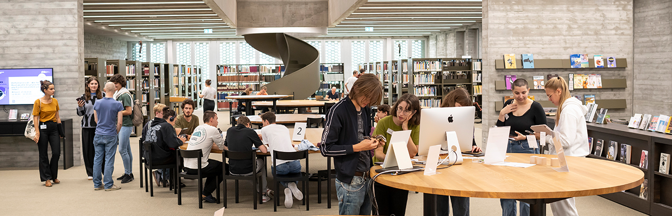 Menschen stehen an einem grossen runden Tisch in der Bibliothek, im Hintergrund sitzen weitere Menschen