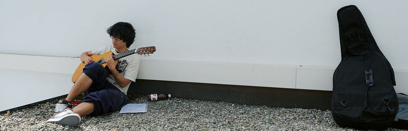 Student lehnt sich an einer Wand und spielt sitzend Gitarre.