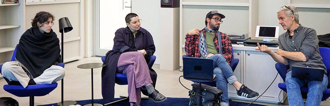 Drei Studierende sitzen auf blauen Sesseln un lassen sich etwas vom Dozent erklären