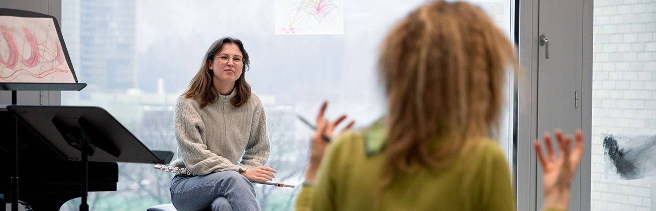 Eine Dozentin, die der Kamera den Rücken zukehrt und  nicht fokussiert ist, erklärt einer aufmerksamen Querflöten-Studentin etwas