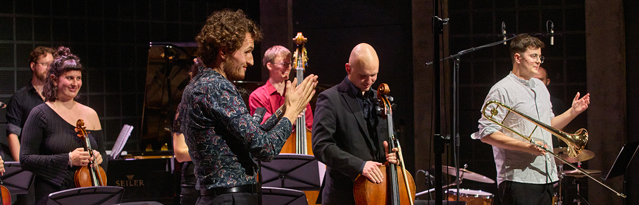 Musiker*innen auf der Bühne, Geige, Bass und Cello, ein Musiker applaudiert