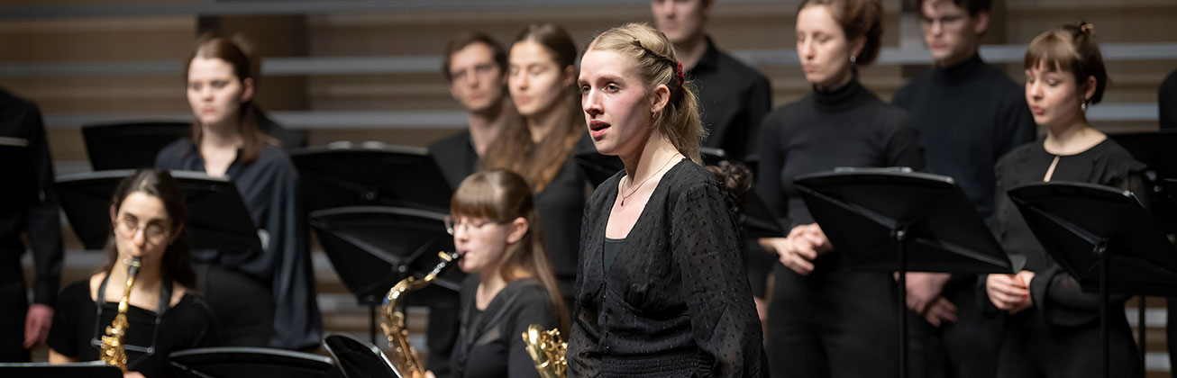 Eine junge Frau singt auf der Bühne, im Hintergrund sitzen Musikerinnen mit Saxophonen und weitere Chormitglieder stehen an Notenständern.