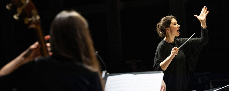 Eine junge Dirigentin vor schwarzem Hintergrund leitet ein Ensemble.