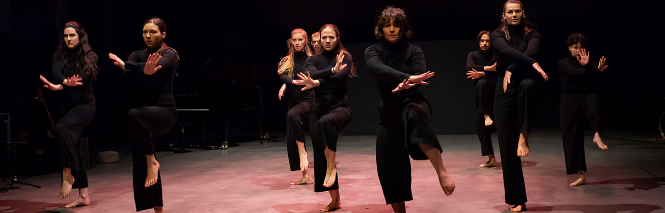In Schwarz gekleidete Studentinnen performen einen Tanz auf der Bühne in rötlichem Licht, der Hintergrund ist Schwarz